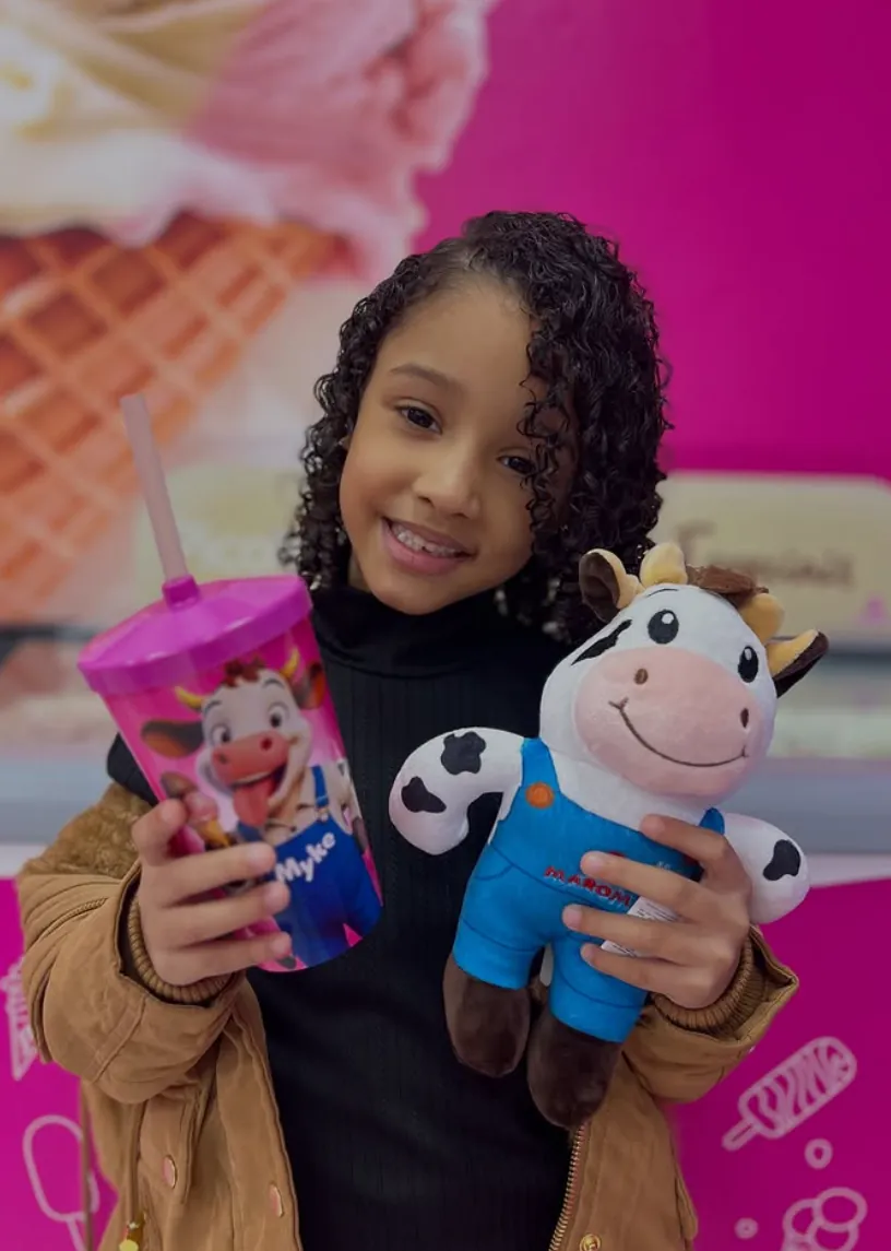 Criança sorrindo com copo Maroma e pelúcia do mascote Myke